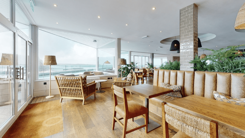 A screenshot from a virtual tour of a hotel lobby and restaurant. The image shows restaurant tables and large windows with a view of the sea